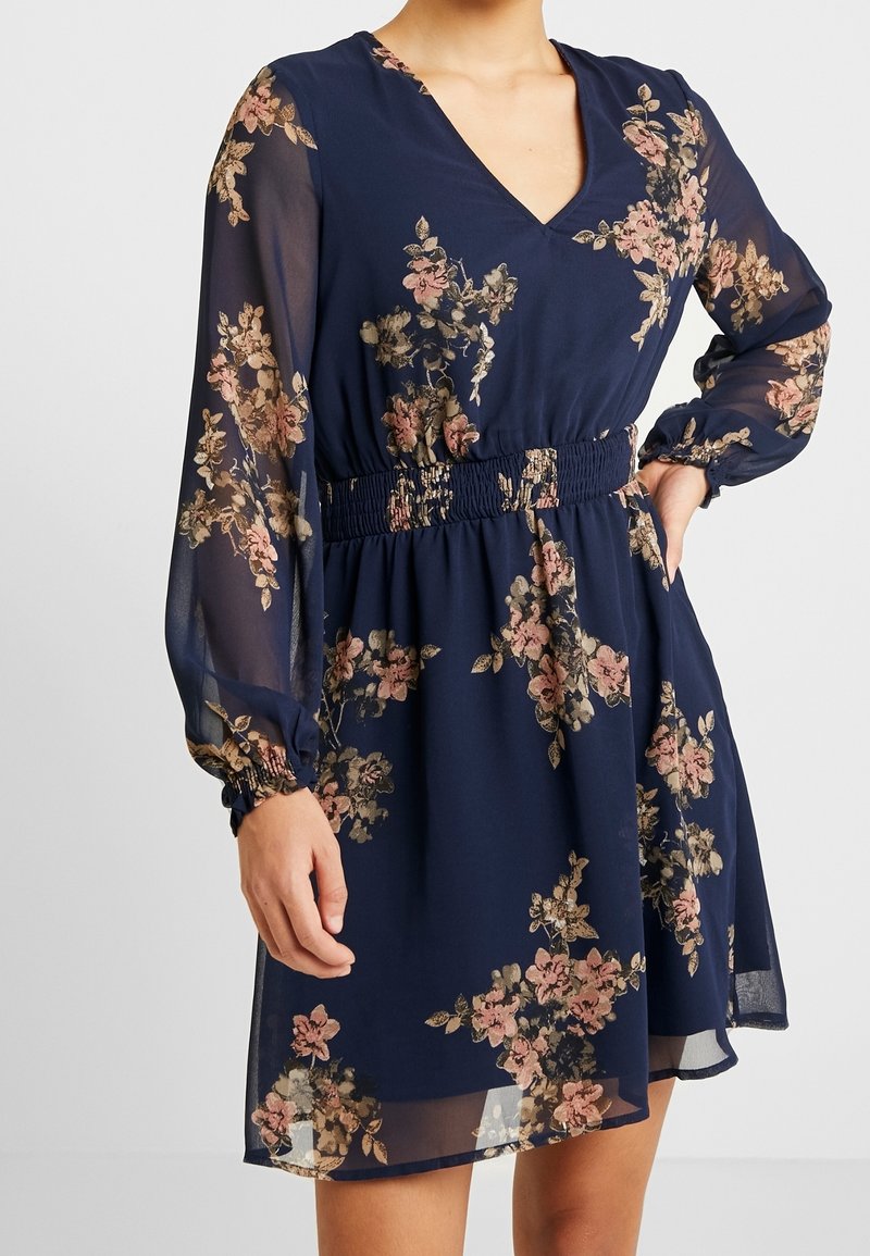 Femme portant une robe bleu marine à fleurs avec des manches longues transparentes, un décolleté en V et une taille smockée, debout devant un fond uni.
