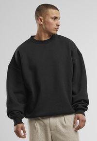 Felpa nera oversize in cotone morbido, con scollo rotondo, spalle scese e polsini a coste. Abbinata a pantaloni beige.