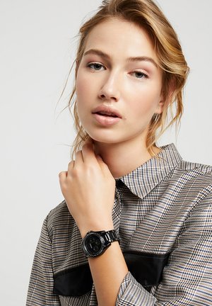 Jeune femme aux cheveux clairs portant une chemise à carreaux et une montre-bracelet noire, tenant sa main près de son cou sur un fond uni.