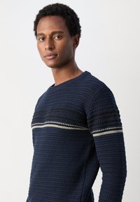 Mørkeblå strikkegenser med et teksturert mønster, med en horisontal stripe av sølv og svart på brystet. Rund hals.