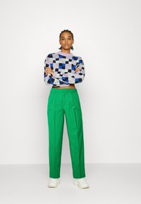 Pulôver de malha com padrão de quadros em roxo, azul e preto; calças verdes de cintura alta; tênis brancos; braços cruzados, fundo neutro.