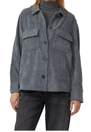 Kobieta w szarej sztruksowej kurtce zapinanej na guziki z klapkowymi kieszeniami na piersi, pod którą ma szary golf, oraz czarne dżinsy.