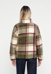 Zöld, krémszínű és burgundi kockás pléddzseki; magas nyak, oversized szabás és texturált anyag, amely kockás mintával rendelkezik.