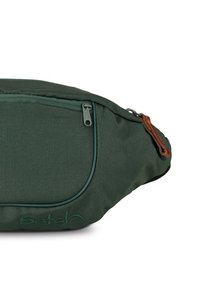 Grüne Stoff-Tašche mit einem vorderen Reißverschlussfach, lederner Reißverschlusslasche und Logostickerei. Glatte Textur und geschwungenes Design.