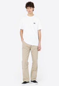 White short-sleeve t-shirt featuring a small graphic logo, paired with light beige straight-leg pants and black slip-on shoes.