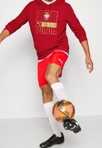 Felpa rossa con stampa e logo dorati, pantaloni corti rossi, calze bianche, scarpe da calcio nere e un pallone da calcio arancione. Messa a fuoco su un design sportivo e atletico.