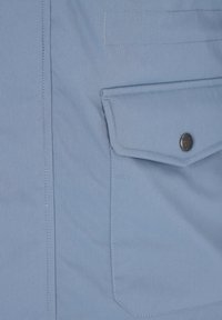 Tela azul claro con un bolsillo de solapa rectangular asegurado por un botón a presión de metal oscuro, con costuras detalladas a lo largo de los bordes.