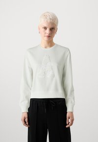 Hanorac verde deschis cu un design texturat alb în formă de "A" pe față, guler rotund cu nervuri și manșete ajustate, combinat cu pantaloni negri.
