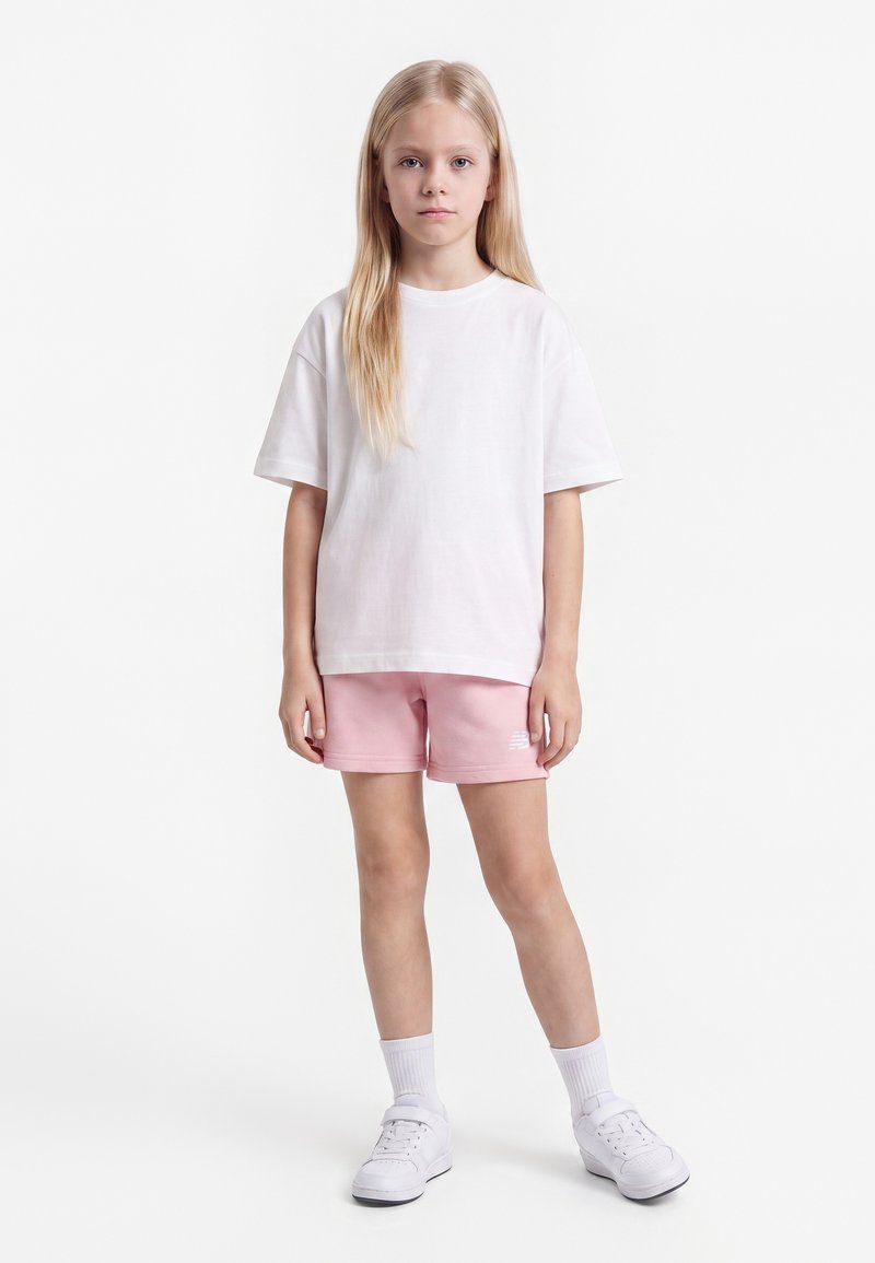 Fille blonde debout, face à l’objectif, portant un t-shirt blanc oversize, un short rose, des chaussettes blanches et des baskets blanches sur fond blanc uni.