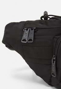 Bolsa preta em nylon com fechos duplos, detalhes em fita e forma arredondada. Tecido texturizado com detalhe de costura. Design compacto.