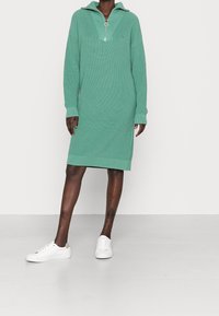 Robe en maille côtelée vert menthe avec col zippé mi-long et fente latérale. Présente des manches longues et une coupe décontractée, associée à des baskets blanches.