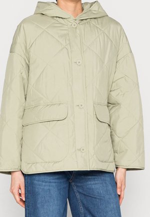 Veste matelassée à capuche beige clair avec boutons sur le devant et poches à rabat, portée avec un jean bleu.