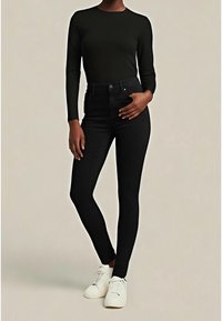 Personne portant un haut noir ajusté à manches longues, un jean skinny noir taille haute et des baskets blanches, debout avec une main sur la hanche.