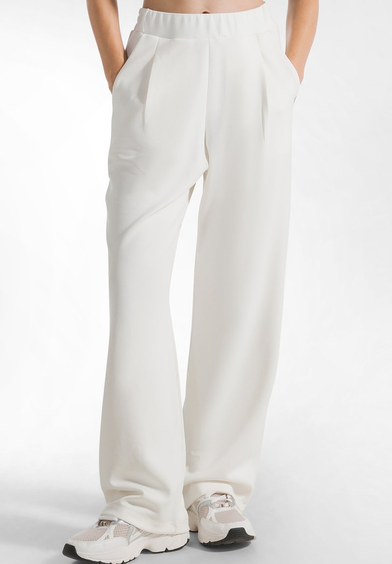 Weiße weit geschnittene Hose mit gefaltetem Vorderteil und elastischem Bund, kombiniert mit hellen Sneakers. Glattes Material, lässiges Design.