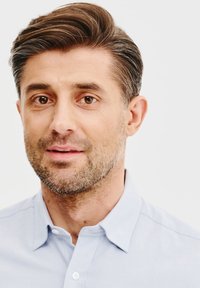 Zbliżenie mężczyzny z brązowymi włosami i jasnoniebieską koszulą z kołnierzykiem. Ma krótką brodę i patrzy bezpośrednio w kamerę na tle białego tła.