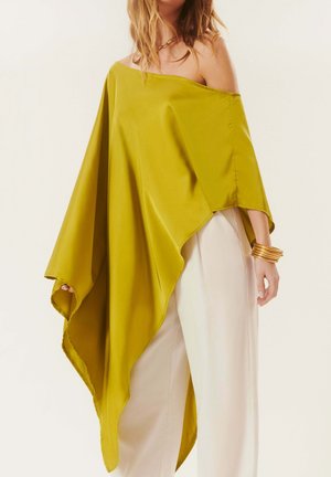 Femme portant un poncho asymétrique vert olive décolleté, un pantalon blanc à jambes larges et des bracelets dorés au poignet.