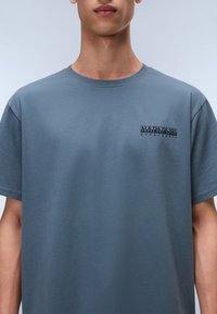 Grå bomuld T-shirt med rund halsudskæring, korte ærmer og et lille sort logo på øverste venstre brystområde. Glat tekstur.