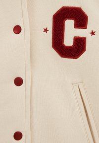 Krämfärgad varsityjacka med en fluffig röd bokstav "C" och matchande runda knappar. Inkluderar sidofickor och textil med struktur.