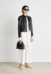 Svart läderjacka, vit topp, vida vita byxor, gula spetsiga ballerinaskor, håller en liten svart handväska, överdimensionerade svarta solglasögon, minimalistisk design.