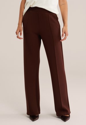 Femme portant un pantalon marron foncé taille haute à jambes larges, associé à un haut blanc et des chaussures pointues foncées, debout sur un fond clair.