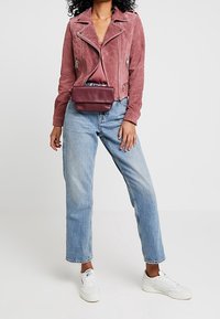 Różowa zamszowa kurtka motocyklowa z srebrnymi zamkami, zestawiona z jasnoniebieskimi prostymi spodniami dżinsowymi i białymi sneakersami. Mała bordowa torebka na ramię.