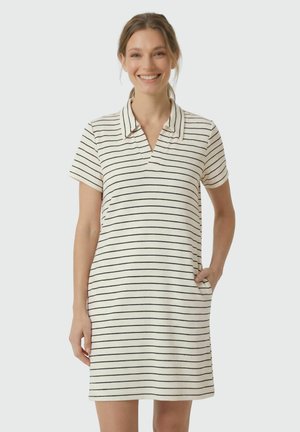 Lächelnde Frau in einem weißen, kurzärmeligen, knielangen Kleid mit schwarzen horizontalen Streifen und Kragen, stehend mit einer Hand in der Tasche.