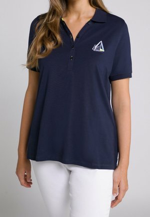 Granatowa koszulka polo z krótkimi rękawami i plisą na trzy guziki. Ozdobiona haftowanym motywem żaglówki. W zestawie z białymi spodniami.