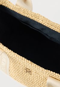 Geflochtener Strohtasche mit beigen Stoffgriffen, offenem Oberteil, das ein dunkles Innenfutter zeigt, mit einem metallischen Logo-Akzent an der Seite.