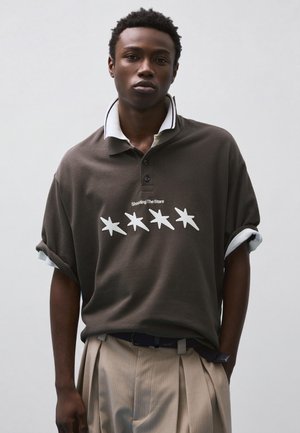 Mladý muž oblečený v tmavom polokošeli s nápisom "Shooting The Stars" a piatimi bielymi hviezdami, v kombinácii s béžovými plisovanými nohavicami.