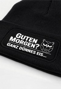 Black knitted beanie featuring a rectangular patch with white text and an owl graphic. Text reads "GUTEN MORGEN? GANZ DÜNNES EIS..."