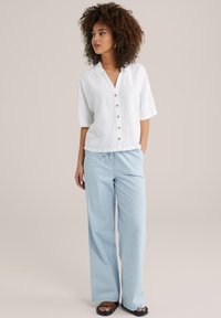 Camicia bianca in lino con bottoni e maniche corte, abbinata a pantaloni larghi a righe blu chiaro. Modella che indossa sandali neri, in piedi su uno sfondo neutro.