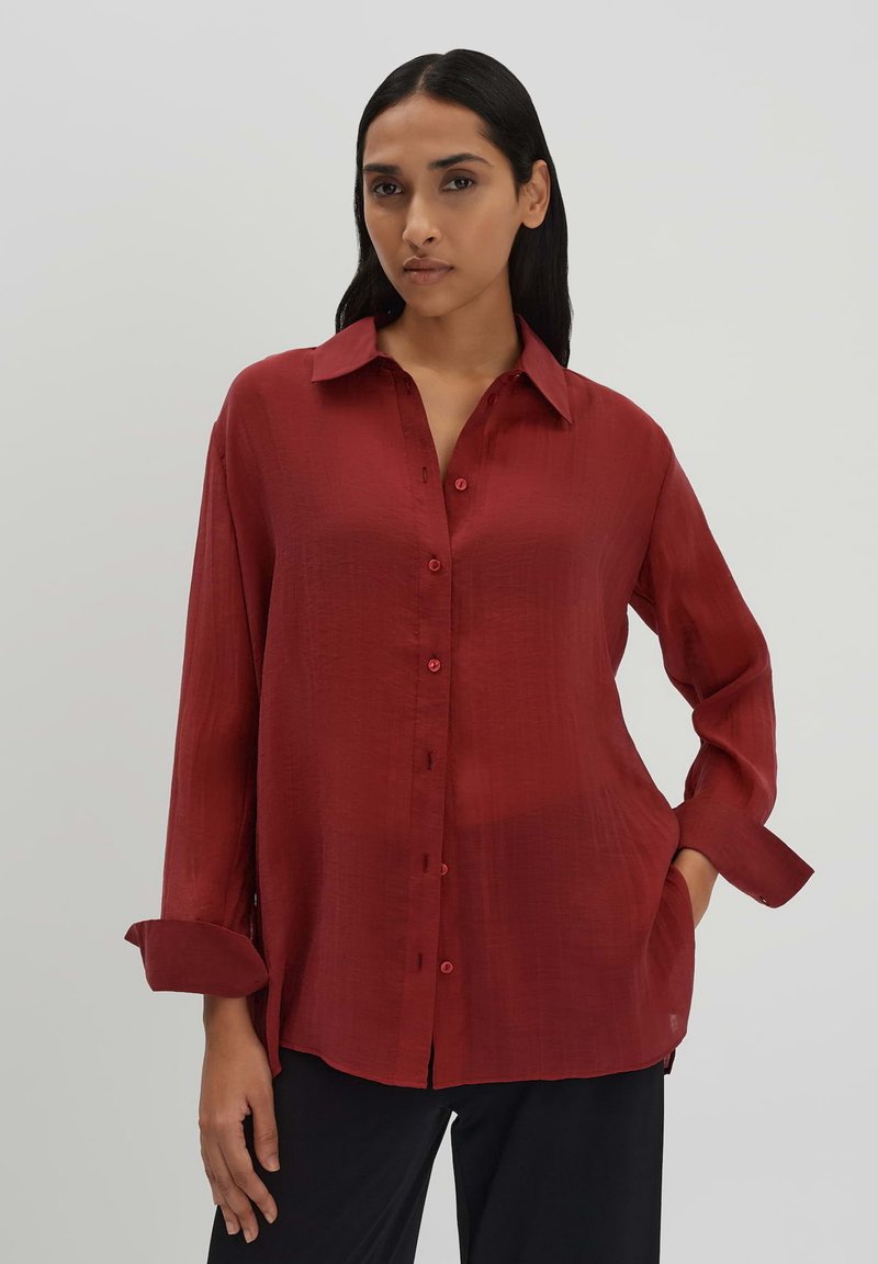 Camicia rossa con bottoni realizzata in tessuto testurizzato, dotata di colletto a punta, maniche lunghe arrotolate ai polsini e fessure laterali per maggiore comfort.
