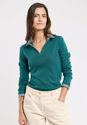 Vrouw in een teal kleurige longsleeve shirt met een bloemenkraag en manchetten, beige broek, staand met één hand in haar zak tegen een lichte achtergrond.