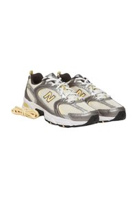 Zapatillas deportivas con parte superior de malla en gris y beige con detalles amarillos, suela resistente, puntera redondeada y cordones blancos.