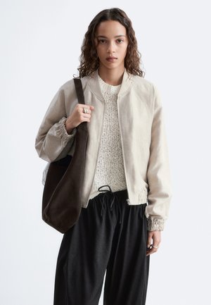 Young woman with curly hair wearing beige jacket, cream knit sweater, black pants, and carrying a large dark brown shoulder bag.