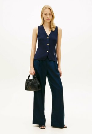 Donna che indossa un gilet blu navy senza maniche con bottoni e pantaloni a gamba larga, che tiene una borsa nera con manici annodati, in piedi su uno sfondo bianco.