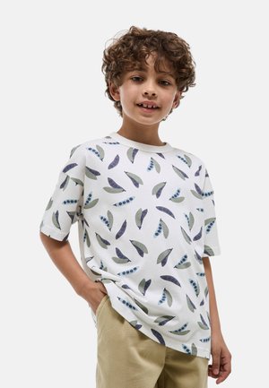 Enfant aux cheveux bouclés portant un t-shirt blanc avec un motif de cosses de pois bleu et vert et un pantalon beige, debout avec une main dans la poche.