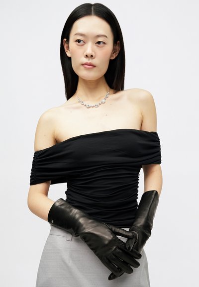 Moteris su tiesiais tamsiais plaukais vilki juodą palaidinę be pečių, sidabrinę gėlių kaklo puošmeną, ilgas juodas odines pirštines ir šviesiai pilkus kelnes.