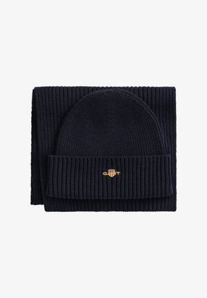Ensemble bonnet et écharpe en tricot bleu marine, présentant une texture côtelée et un design plié, avec un petit logo brodé sur le bonnet.