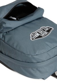 Szary plecak z materiału z klapką na górze, z łatką z logo Vans w czarno-białych kolorach i kieszenią na zamek. Teksturowana powierzchnia.