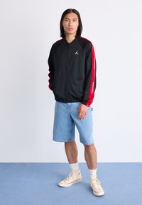 Black zip-up jacket with red accents and Jordan logo, paired with light blue denim shorts and beige high-top sneakers.