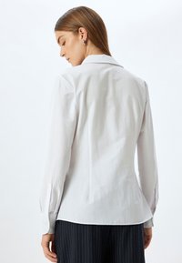 Camicia bianca a manica lunga con colletto classico, vestibilità tailored e polsini abbottonati. Tessuto liscio e leggermente lucido con pince posteriori per una forma definita.