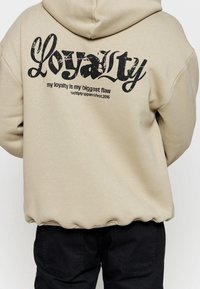 Beige mikina s kapucí s velkým černým potiskem na zádech, který říká "Loyalty" a sloganem. Dlouhé rukávy s volným střihem a žebrovaným lemem.