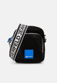 Črn najlonov crossbody nahrbtnik z modrim logotipom, prirejenim trakovi z znakom, zadrgo za zapiranje in kompaktno pravokotno obliko.