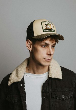 Joven mirando hacia la derecha, lleva una gorra beige y negra con un parche que dice "Beaver Dept Timber Patrol", una chaqueta negra con cuello de borrego y una camisa blanca.