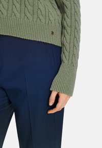 Grüner Kabelstrickpullover mit gerippten Bündchen, ausgestattet mit einem kleinen Metall-Logo. Über dunkelblauem, maßgeschneidertem Hosen getragen, was eine glatte Textur zeigt.