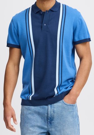 Homme portant un polo bleu à manches courtes avec des rayures verticales bleu clair et bleu foncé, associé à un jean denim bleu clair, une main dans la poche.