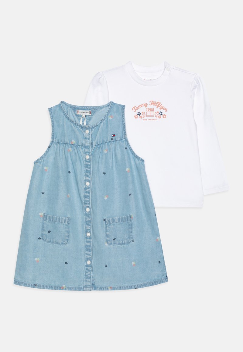 Vestito senza maniche in denim con ricami floreali, due tasche frontali, chiusura a bottoni; abbinato a una camicia bianca a maniche lunghe con testo stampato.