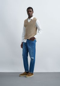 Mann steht mit der Hand in der Tasche, trägt eine beiges Strickweste über weißem Hemd, blaue zerrissene Jeans und braune Sneaker vor einfachem Hintergrund.