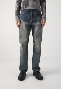 Light blue denim jeans with a relaxed fit, featuring fading and whiskering details. Paired with black leather shoes and a patterned grey top.